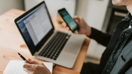 En la foto: Hombre con celular en la mano, de fondo un computador portátil.